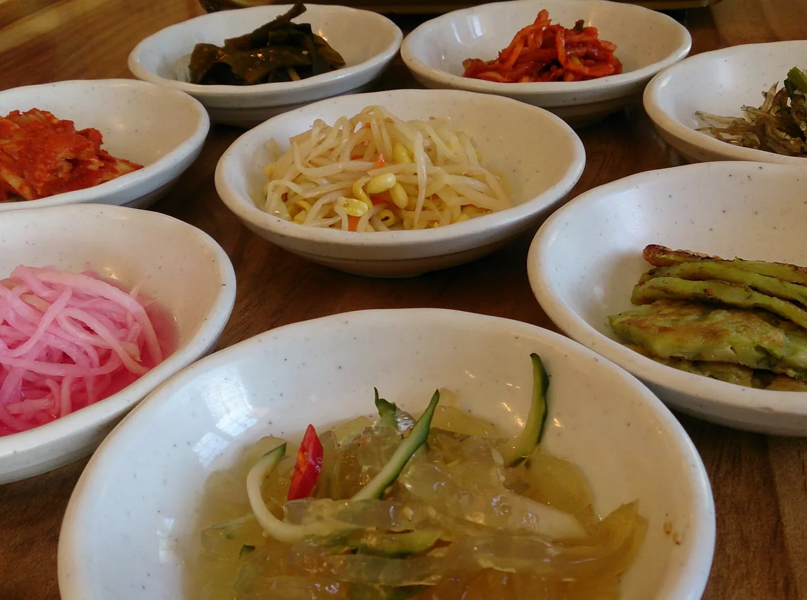 White bowls of banchan on a wooden table, among them red kimchi, bean sprouts, pink pickled radish and cucumber in a clear chilled broth