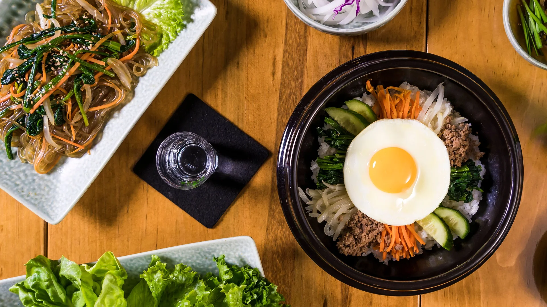 Overhead view of a black stone bowl of bibimbap topped with a fried egg, carrot, spinach, bean sprouts and minced beef on a wooden table, with a plate of japchae glass noodles beside it