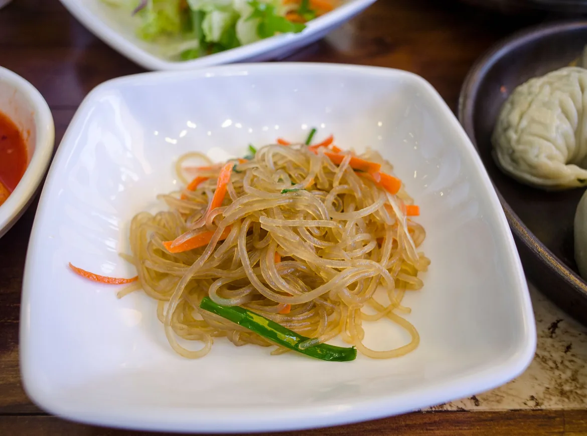 A white square plate of japchae, glass noodles tossed with carrot and spring onion, with a dumpling and small side dishes beside it