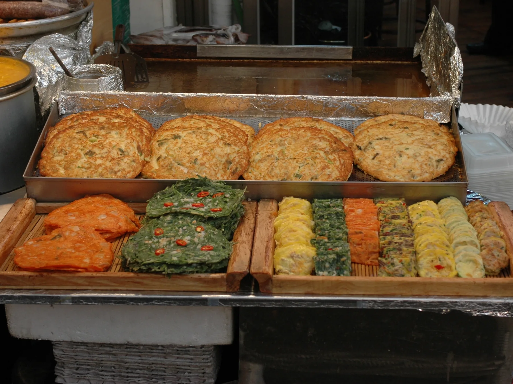 Trays of Korean pan-fried pancakes at a Gwangjang Market stall: four large golden rounds above rows of green, orange and yellow jeon
