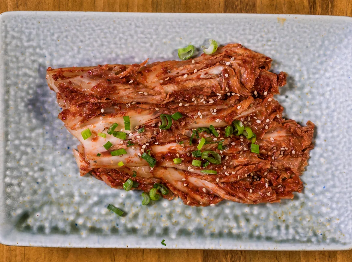 A halved leaf of napa cabbage kimchi dressed in red chilli paste and sprinkled with sesame seeds and spring onion on a pale blue rectangular plate