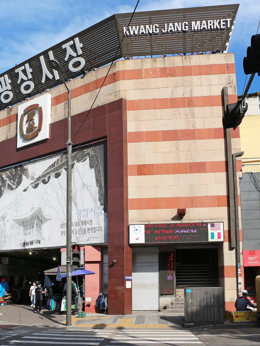 The red-and-cream striped entrance building of Gwangjang Market in Seoul under a blue sky, with the market’s name in Korean and English on the roof sign